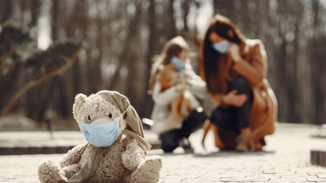 Cute children toy in scarf and mask placed on asphalt against blurred owners woman and girl in warm wear playing in nature