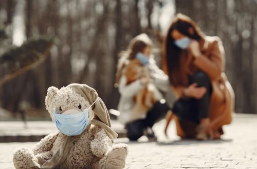 Cute children toy in scarf and mask placed on asphalt against blurred owners woman and girl in warm wear playing in nature