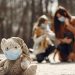 Cute children toy in scarf and mask placed on asphalt against blurred owners woman and girl in warm wear playing in nature